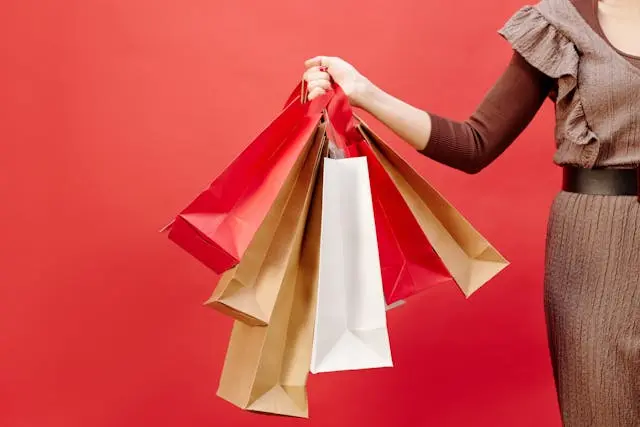 Người phụ nữ mặc váy nâu đang cầm trên tay nhiều chiếc túi giấy mua sắm màu nâu, trắng và đỏ trên nền đỏ trơn nổi bật.